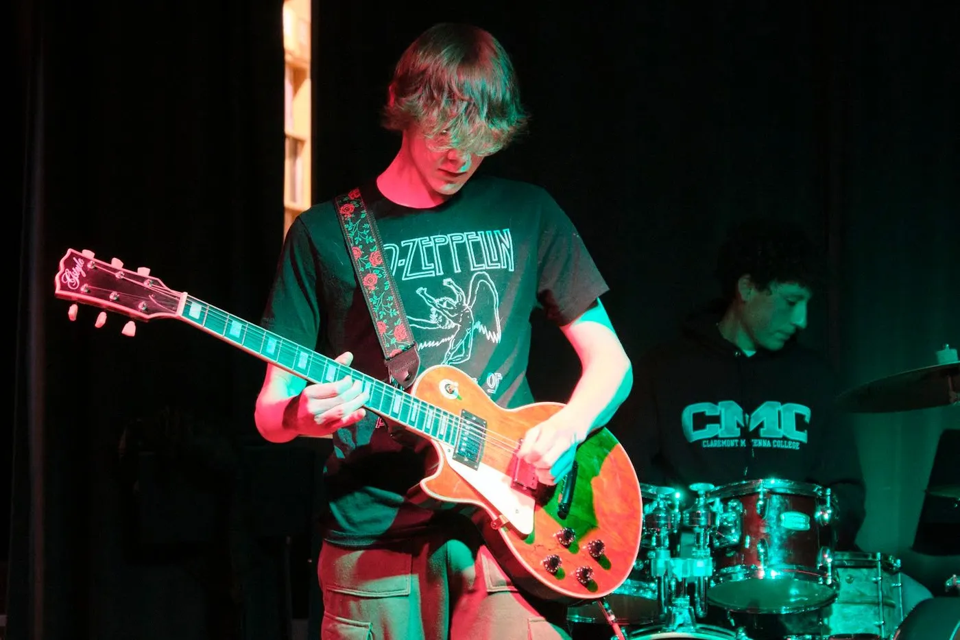 A teen guitarist plays an electric guitar on stage with a drummer behind him