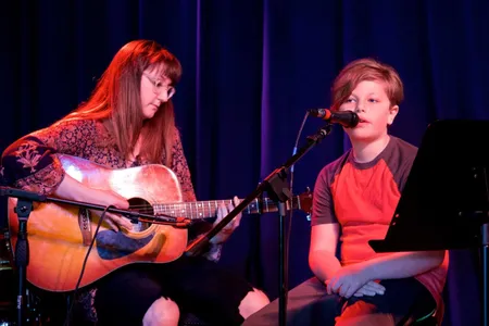 A young singer performs onstage beside his guitar teacher