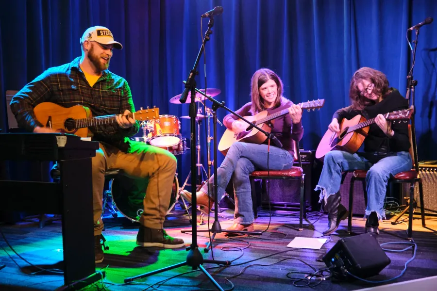 A guitar teacher and two students play acoustic guitars together on stage