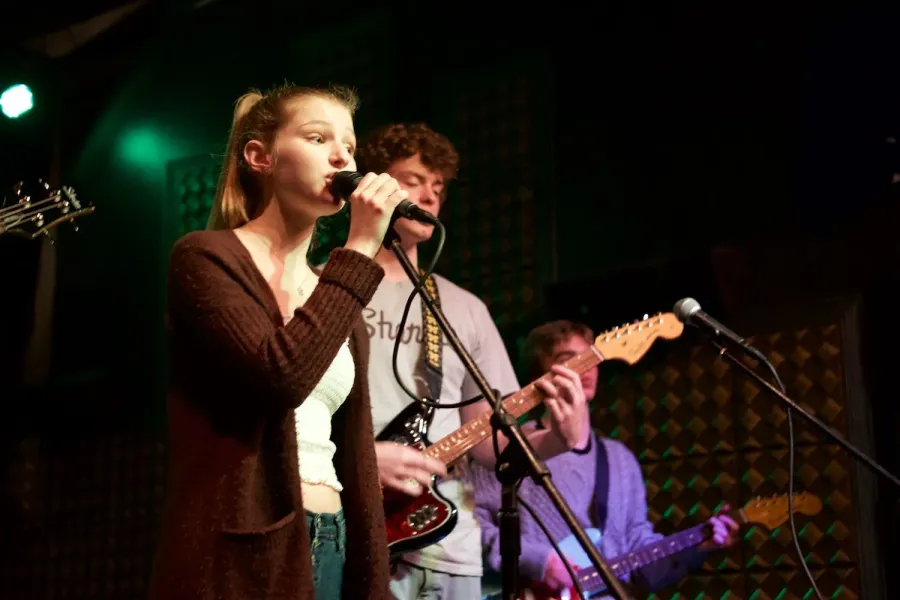 A student sings lead into the microphone while two bandmates play guitar behind her