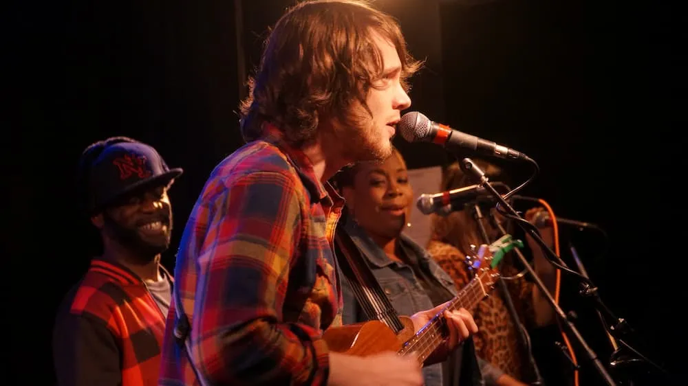 A musician sings and plays ukulele on stage at a Sunburst open mic